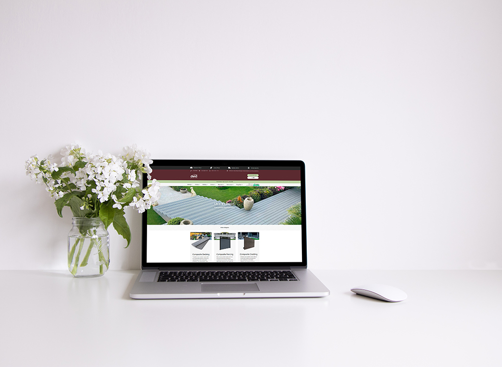 Laptop displaying a home improvement website with a focus on composite decking and fencing, accompanied by a vase of white flowers on a clean white desk.