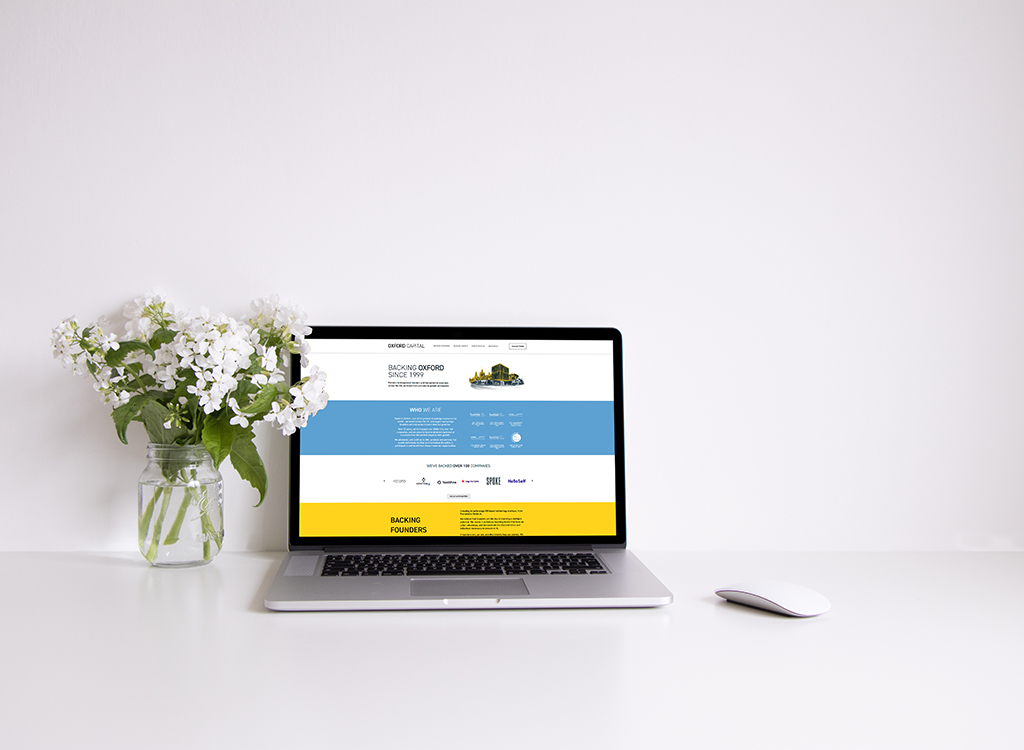 Laptop displaying the Oxford Capital website, featuring a clean design with sections on company information and backing founders, set on a minimalist white desk alongside a glass vase of white flowers.