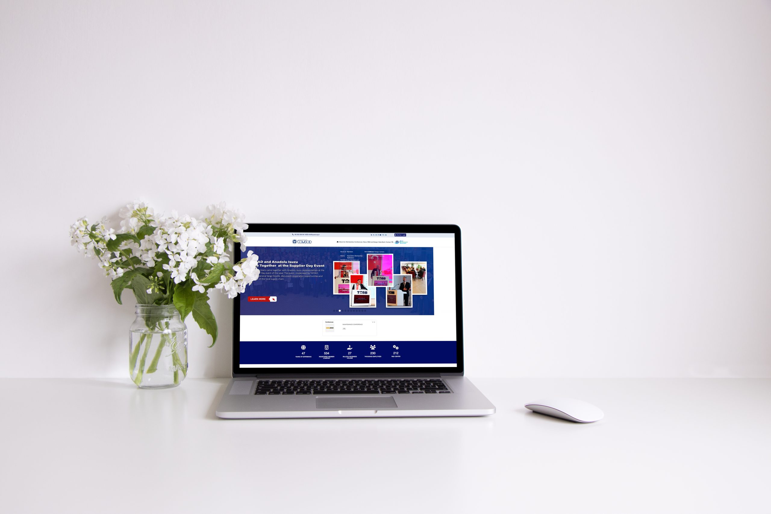 A modern workspace featuring a laptop displaying a website, accompanied by a vase of white flowers and a computer mouse, set against a minimalist white background.