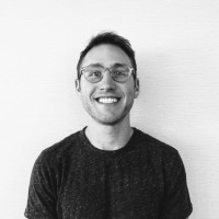 Smiling young man with glasses wearing a dark t-shirt, standing against a light textured background.