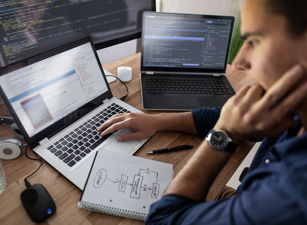 A focused software developer coding on a laptop, surrounded by two additional screens displaying programming code, with notes and flowcharts on a notepad, highlighting a work environment for web development or software engineering.