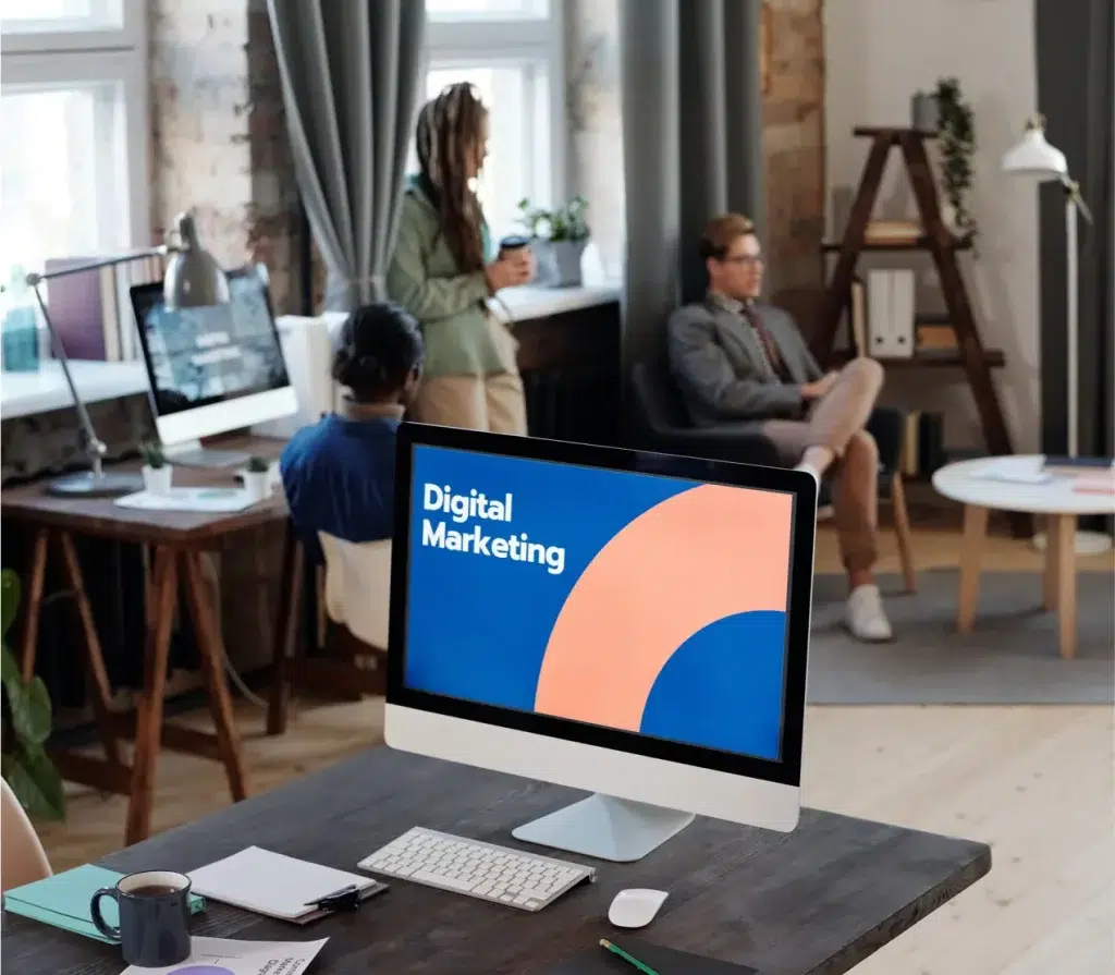 A modern office setting featuring a desktop computer displaying "Digital Marketing" on the screen. In the background, a woman stands holding a coffee cup, while two colleagues engage in conversation. The workspace includes stylish furniture, large windows, and a relaxed atmosphere, emphasizing creativity and collaboration in digital marketing.